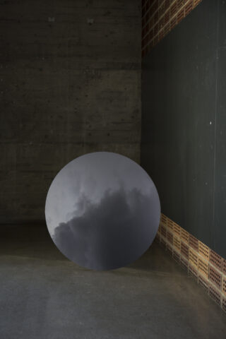 Ein großer runder Spiegel lehnt an einer dunklen Wand und reflektiert einen bewölkten Himmel. Die Kulisse ist ein Industriegebäude mit Betonböden und -wänden.