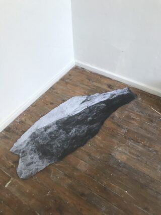Ein Schwarzweißdruck eines Felsens liegt flach auf einem Holzboden in der Ecke eines Raums mit weißen Wänden.