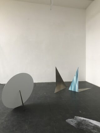Minimalistische Kunstinstallation mit drei geometrischen Metallskulpturen: einer runden Scheibe, einem flachen silbernen Dreieck und einem silbernen Dreieck mit himmelblauem Muster, platziert auf einem dunklen Fliesenboden.