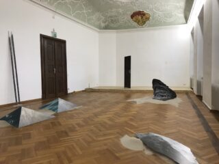 Eine Kunstinstallation in einem Raum mit dekorativer Decke, Holzboden, abstrakten Skulpturen, die an Steine und Sand erinnern, und einer geschlossenen Doppeltür.