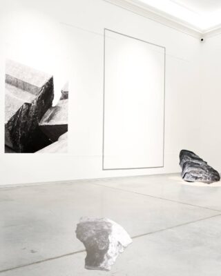 Ein minimalistischer Galerieraum mit einem großen Schwarzweißfoto an der Wand, zwei Steinen auf dem Boden und einem schwachen rechteckigen Umriss an der Wand.