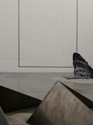 Eine minimalistische Kunstinstallation für den Innenbereich mit eckigen, mit Sand gefüllten Metallstrukturen und einem großen, dunklen Felsen vor einer schlichten Wand mit geometrischen Liniendetails.
