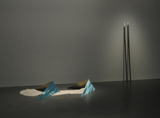 Eine minimalistische Kunstinstallation besteht aus zwei blauen dreieckigen Glasstrukturen und zwei braunen Formen, die teilweise im Sand vergraben sind, wobei zwei hohe schwarze Stangen in einem schwach beleuchteten Raum an einer grauen Wand lehnen.