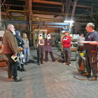 Eine Gruppe von Menschen steht in einer Werkstatt, einige halten rechteckige Gegenstände in den Händen, während ein Mann in Arbeitskleidung mit ihnen zu sprechen scheint.