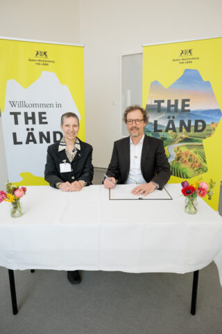 Zwei Personen sitzen an einem Tisch und unterschreiben Dokumente. Im Hintergrund sind Banner mit der Aufschrift „Das Land“ zu sehen. Vorne auf dem Tisch steht eine Vase mit Blumen.
