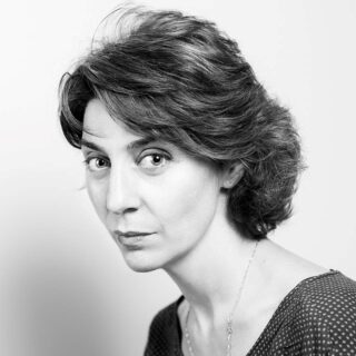 Schwarz-Weiß-Porträt einer Frau mit kurzem, welligem Haar, die ein gemustertes Oberteil trägt und mit neutralem Gesichtsausdruck direkt in die Kamera blickt.