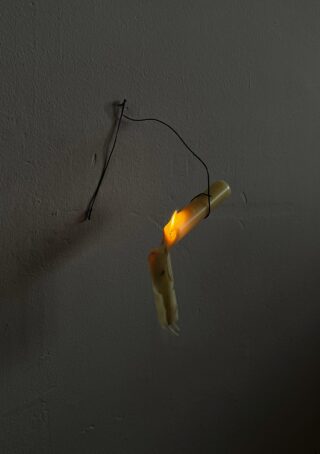 Eine brennende, teilweise geschmolzene Kerze hängt an einem gebogenen Draht, der an einer schlichten Wand befestigt ist, und wirft ein schwaches Licht und einen kleinen Schatten.