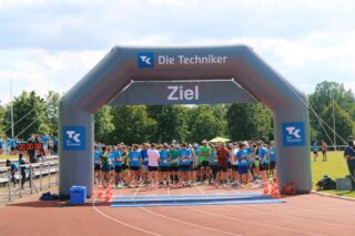 Eine Gruppe von Läufern in blauen Shirts versammelt sich bei einem Laufwettbewerb im Freien an der Startlinie unter einem aufblasbaren Bogen mit der Aufschrift „Ziel“.
