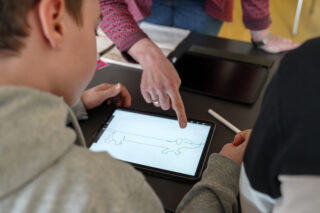 Zwei Kinder schauen auf den Bildschirm eines Tablets, auf dem eine Hundezeichnung zu sehen ist, während ein Erwachsener auf den Bildschirm zeigt und ein Stift in der Nähe auf dem Tisch liegt.