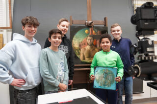 Fünf Jungen stehen lächelnd in einem Atelier vor einer Staffelei und halten ein Gemälde eines Mannes in den Händen. Ein Junge hält ein kleineres Kunstwerk in den Händen. Im Vordergrund ist eine Kamera aufgestellt.