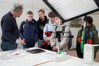 Ein Erwachsener erklärt einer Gruppe von fünf Schülern in einem Klassenzimmer mit wissenschaftlichen Geräten und Displaylichtern einen Gegenstand auf einem Tisch.