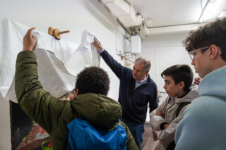 Ein Mann hebt ein Blatt an, um drei Schülern in einem Klassenzimmer oder einer Werkstatt ein Kunstwerk zu zeigen.