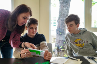 Eine Frau hilft zwei Jungen an einem Tisch mit einem grünen 3D-gedruckten Objekt, während ein anderer Junge aufmerksam zusieht.