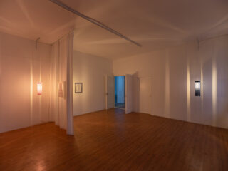 Minimalistische Kunstgalerie mit Holzböden, weißen Wänden, Stofftrennwand, gerahmter Kunst, zwei Hängelampen und offenen Doppeltüren, die den Blick auf einen blau beleuchteten Nebenraum freigeben.