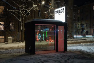 Ein kleiner Glaskiosk mit der Aufschrift „DISCO“ ist nachts beleuchtet und zeigt im Inneren bunte Neonlichter, umgeben von Schnee auf dem Boden und nahe gelegenen Gebäuden.