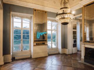 Eleganter, historischer Raum mit Parkettboden, Kronleuchter, großen Fenstern mit Blick aufs Wasser und einem blauen Neonlicht-Kunstwerk an der Wand.