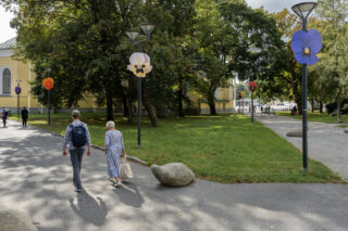 Zwei Menschen gehen einen Weg in einem Park mit grünem Gras und Bäumen entlang; Laternenpfähle sind mit großen künstlichen Blumen geschmückt.