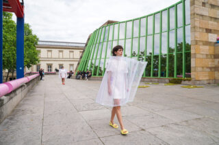 Eine Person in einem weißen Kleid und einem durchsichtigen Plastikumhang geht auf einem gepflasterten Platz vor einem modernen Gebäude mit grün gerahmten Fenstern.