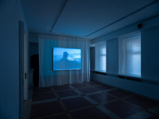 Ein schwach beleuchteter Raum mit einem großen Bildschirm, auf dem die Silhouette einer Person vor statischem blauem Licht zu sehen ist, umgeben von weißen Vorhängen und zwei hohen Fenstern.