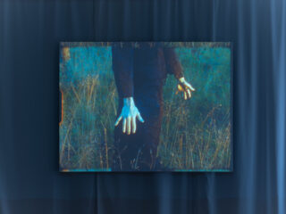 Auf einem digitalen Bildschirm ist das Bild einer Person in dunkler Kleidung zu sehen, die auf einer Wiese steht. Nur ihr Oberkörper und ihre blassen Hände sind zu sehen.