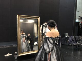 Eine Frau in einem schwarz-weißen historischen Kostüm steht hinter der Bühne vor einem großen Spiegel. Ihr Spiegelbild ist zusammen mit dem einer weiteren Person neben ihr sichtbar.