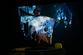 Eine Person steht vor einem projizierten Bild von Bäumen und blauem Himmel, wobei Teile der Projektion auch auf ihren Körper und einen durchscheinenden Stoff projiziert werden, den sie hält.