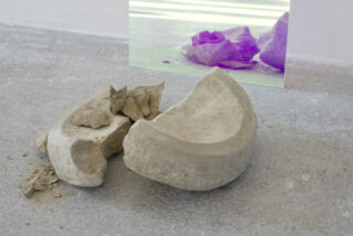 Ein zerbrochener, hohler, steinartiger Gegenstand liegt auf einem Betonboden. In der Nähe sind Fragmente verstreut und im Hintergrund zeigt eine reflektierende Oberfläche einen violett getönten Widerschein.