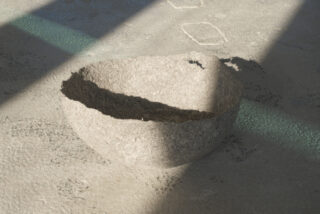 Eine strukturierte, flache, graue Schüssel steht auf einer rauen, sonnenbeschienenen Oberfläche, über die schräge Schatten fallen.