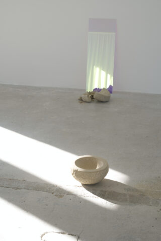 Eine runde, strukturierte beige Schüssel steht auf einem Betonboden. Im Hintergrund lehnen Steine und ein hoher, pastellfarbener Spiegel an einer weißen Wand. Sonnenlicht erzeugt diagonale Schatten.