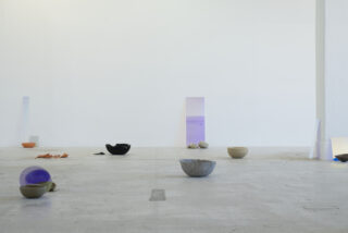In einem minimalistischen Galerieraum mit weißen Wänden sind mehrere Schalen und abstrakte Objekte in verschiedenen Farben auf einem nackten Betonboden angeordnet.