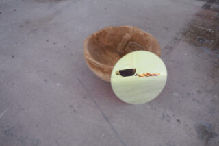 Ein runder Spiegel auf Beton reflektiert eine Holzschale und verstreute Gegenstände, während die eigentliche Holzschale direkt hinter dem Spiegel steht.