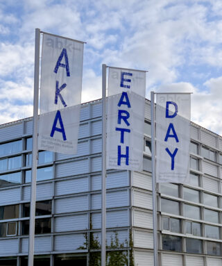 Drei vertikale Banner vor einem modernen Gebäude zeigen die Wörter „AKA“, „EARTH“ und „DAY“ in blauen Buchstaben vor einem bewölkten Himmel.