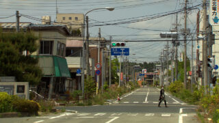Eine Person überquert unter einem teilweise bewölkten Himmel eine leere Straße in einer Kleinstadt mit überwucherten Pflanzen, verlassenen Gebäuden und Freileitungen.