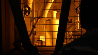 Eine Person beobachtet durch ein schützendes Glasfenster Industrieanlagen mit großen Metallrohren und Maschinen, die von gelb-orangefarbenem Licht angestrahlt werden.