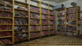 Raum mit gelben Metallregalen, gefüllt mit zahlreichen alten, ungeordneten Akten und Dokumenten; einige sind ordentlich gestapelt, während andere chaotisch und beschädigt wirken.