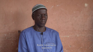 Ein Mann in blauem Hemd und gemusterter Mütze sitzt vor einer strukturierten, rötlichen Wand. An seinem Hemd ist ein Mikrofon befestigt. Der weiße Untertitel lautet: „Rádio Libertação betrieben.“.