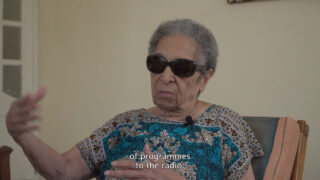 Eine ältere Frau mit dunkler Sonnenbrille und gemustertem Oberteil sitzt auf einem Stuhl und gestikuliert mit einer Hand. Im Untertitel heißt es: „Von Programmen zum Radio.“.