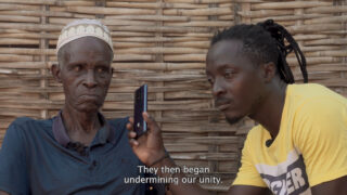 Ein älterer und ein jüngerer Mann sitzen neben einer geflochtenen Wand. Der jüngere Mann hält dem älteren Mann ein Telefon hin. Im Untertitel heißt es: „Dann begannen sie, unsere Einheit zu untergraben.“.