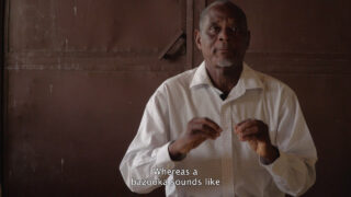 Ein älterer Mann in einem weißen Hemd gestikuliert mit seinen Händen, während er drinnen sitzt; im Untertitel heißt es: „Wohingegen eine Bazooka klingt wie …“.