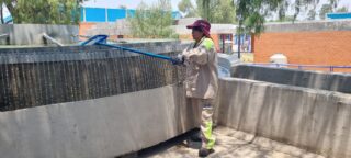 Ein Arbeiter in Schutzkleidung verwendet ein Werkzeug mit langem Griff, um einen großen Industriewassertank im Freien zu reinigen oder zu inspizieren.