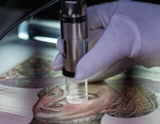 Eine behandschuhte Hand untersucht ein gemaltes Porträt mit einem Vergrößerungswerkzeug und konzentriert sich zur Betrachtung oder Analyse auf das Gesicht.