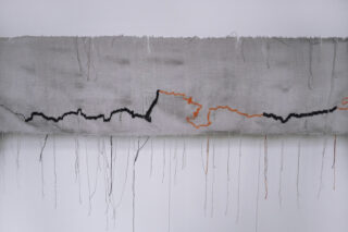 Ein rechteckig gewebter Stoff mit losen vertikalen Fäden und einer gezackten schwarz-orangefarbenen Nahtlinie, die horizontal über den hellgrauen Stoff verläuft.