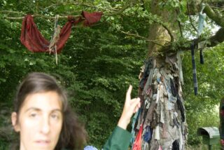 Eine Frau zeigt auf einen Baum in einem Waldgebiet, an dessen Ästen und Stamm viele Stoff- und Kleidungsstücke befestigt sind.