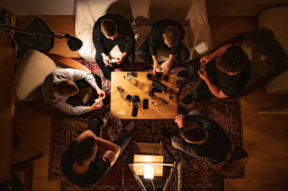 Sechs Personen sitzen in einem Wohnzimmer um einen Tisch herum (von oben betrachtet), auf der Tischplatte stehen Getränke und Gegenstände.