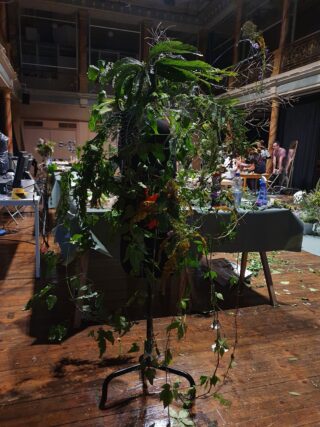 Eine mit verschiedenen grünen Blättern und Pflanzen bedeckte Schaufensterpuppe steht in einem schwach beleuchteten Raum mit Tischen und Menschen im Hintergrund.