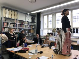 Eine Frau steht in einem markanten Lagenrock auf einem Tisch, während andere, die um den Tisch herum sitzen oder stehen, sie in einem Klassenzimmer oder Studio mit Bücherregalen und Schneiderpuppen beobachten.