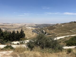 Eine trockene, hügelige Landschaft mit kurvigen Straßen und vereinzeltem Grün unter blauem Himmel; in der Ferne ist eine Stadt zu sehen.