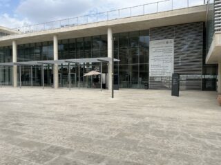 Modernes Gebäude mit Glaswänden, großem Überhang und einem mehrsprachigen Schild auf der rechten Seite; davor leerer Betonplatz.