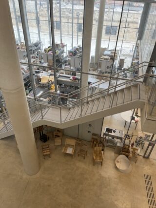 Großer, offener Industriearbeitsbereich mit Tischen, Geräten und Materialien, die durch hohe Glasfenster sichtbar sind; im Vordergrund eine Metalltreppe und gestapelte Holzstühle.