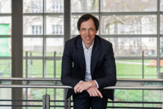 Ein Mann in dunklem Anzug und weißem Hemd lehnt sich nach vorne an ein Geländer in einem Innenraum, in dem große Fenster und eine Grünfläche im Hintergrund zu sehen sind.